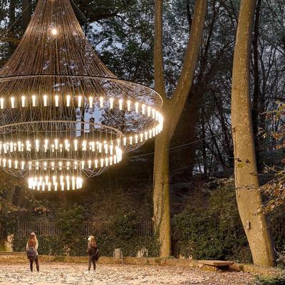 A giant glowing chandelier hanging among trees in a park at dusk